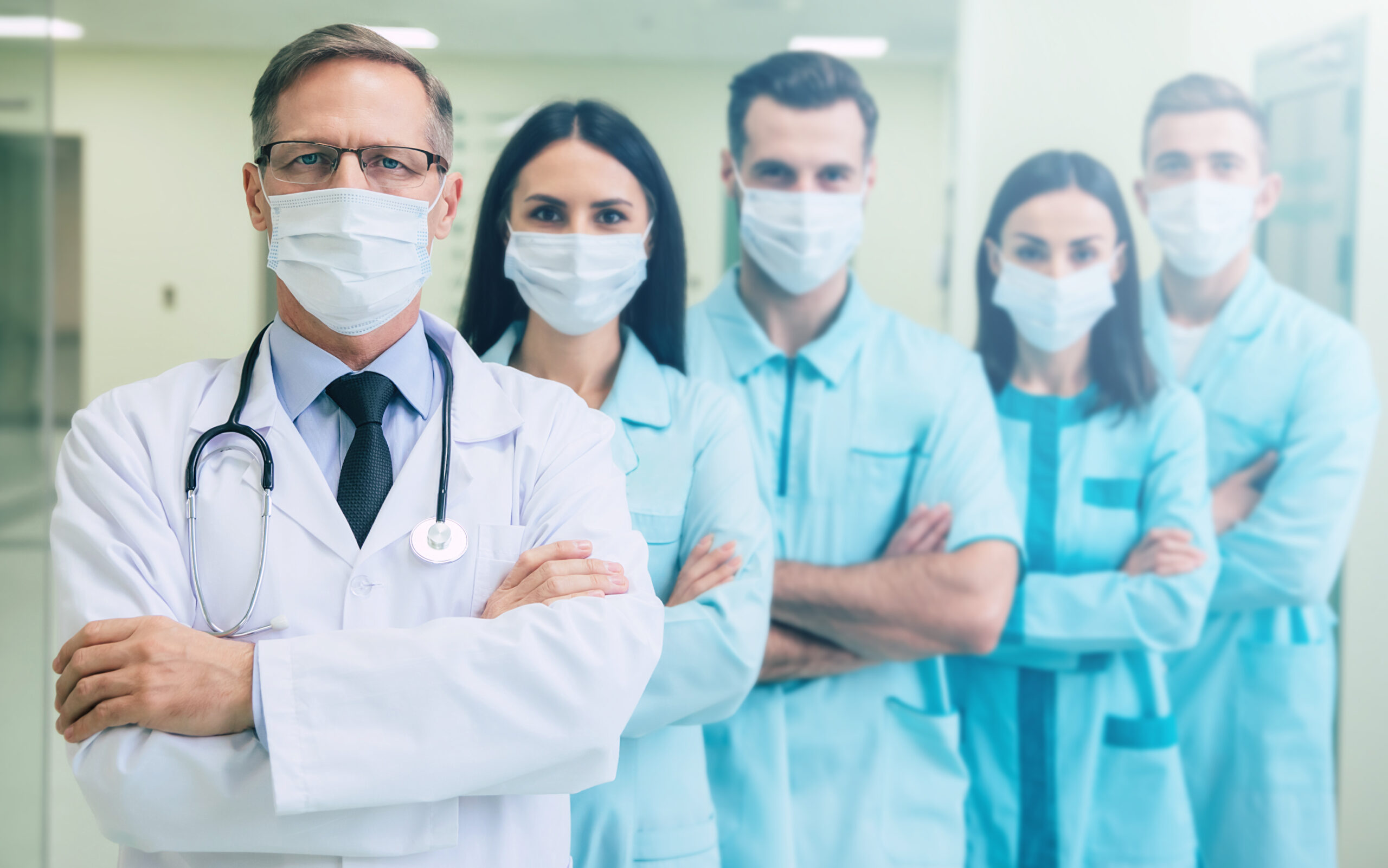 Serious confident doctors team in safety medical masks with crossed arms are looking on the camera on hospital background